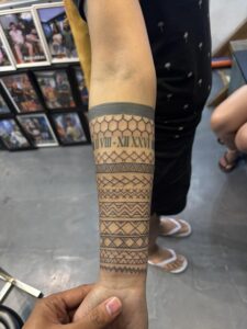 A person's forearm displaying a black ink tattoo with geometric patterns and Roman numerals.