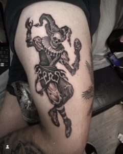 An illustrative black and grey thigh tattoo of a skeletal jester in full costume, wearing a ruffled collar and jester's hat while holding a theatrical mask.