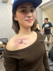 A smiling young woman wearing a blue baseball cap and a brown top, showing a small black tattoo of a stingray on her chest.