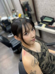 A woman with short, dark hair and bangs looks over her shoulder, showing a red Roman numeral tattoo "MMVI" on her upper shoulder, along with other script tattoos on her arm.