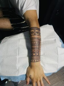 A close-up of a person's arm and hand, showcasing a detailed black tribal armband tattoo that extends from the wrist up the forearm.