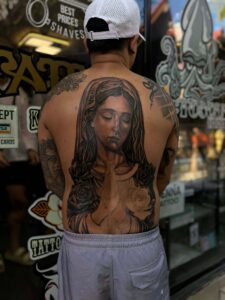 A person with a large tattoo of the Virgin Mary with roses on their back, wearing a white hat and grey shorts.