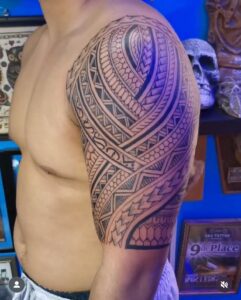 A man's upper arm and shoulder featuring a detailed Polynesian tribal tattoo half-sleeve, incorporating traditional spearhead, shark teeth, and ocean wave patterns in bold black ink.