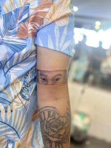 A person's arm with a tattoo of eyes within a rectangular band around the upper arm, and a rose and clock tattoo further down the forearm.