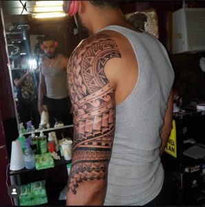 A black ink Polynesian-style tribal tattoo sleeve for a man, featuring intricate geometric patterns, repetitive chevron motifs, and wave patterns, shown in a mirror reflection.