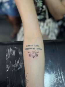 A custom fine-line tattoo on a client’s inner arm by Kurt Bayeta, featuring the Tagalog script 'mahal kita magpakailanman' above a minimalist Philippine Sun with jasmine flowers.
