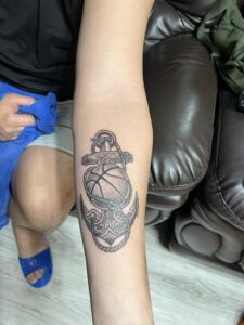 A custom black and grey forearm tattoo by Kurt Bayeta, featuring a detailed basketball integrated with an ornate anchor and nautical rope, showcasing crisp linework and smooth shading.