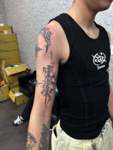 A black and grey illustrative tattoo of a rifle on a person's upper arm, featuring smoking barrel details and falling bullet casings.