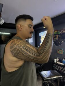 A man with a mustache and a grey tank top raises his arm, proudly displaying a traditional tribal tattoo that covers his forearm and extends towards his elbow.