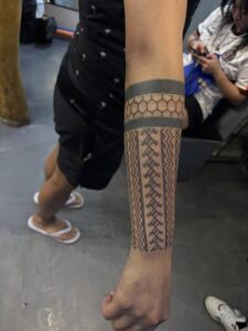 A black ink forearm tattoo featuring a geometric honeycomb pattern framed by bold black bands, transitioning into vertical rows of intricate tribal-style zig-zag and chevron patterns.