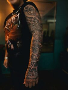 Full-arm realism sleeve tattoo in black and grey, featuring a stairway to heaven with a dove, a boombox with money, and the word 'Believe' on the hand.