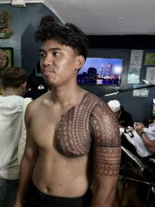 A powerful black ink tribal tattoo covering the chest, shoulder, and upper arm, featuring traditional Polynesian patterns, geometric spearhead motifs, and intricate woven linework.