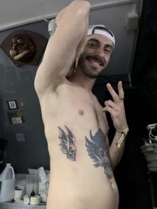 A black and grey side-rib tattoo of a skull with feathered bat wings, featuring clean linework and smooth shading on a male client.