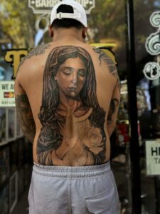 A person with a large tattoo of the Virgin Mary with roses on their back, wearing a white hat and grey shorts.
