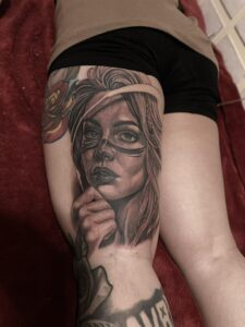 A realistic black and grey thigh tattoo of a woman’s face wearing a decorative lion skull headdress and intricate floral accents, featuring soft shading and lifelike detail.