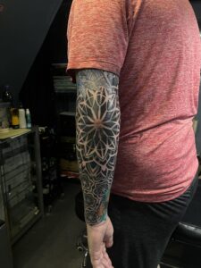 A detailed blackwork geometric tattoo on the elbow featuring a large, symmetrical mandala pattern that extends into an intricate forearm sleeve.