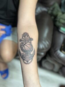 A custom black and grey forearm tattoo by Kurt Bayeta, featuring a detailed basketball integrated with an ornate anchor and nautical rope, showcasing crisp linework and smooth shading.