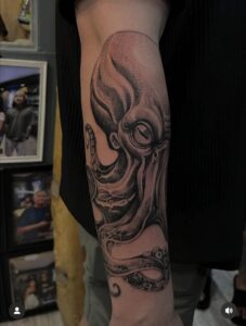 A realistic black and grey forearm tattoo of an octopus, featuring detailed skin texture, prominent suction cups on the tentacles, and deep shading on the head.