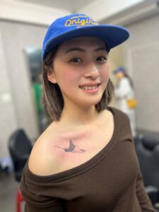 A smiling young woman wearing a blue baseball cap and a brown top, showing a small black tattoo of a stingray on her chest.