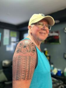 A smiling man with glasses and a tan baseball cap wears a turquoise tank top, gesturing to his arm which features a tattoo of a sun with a palm tree and waves, and a tribal pattern extending down his forearm.