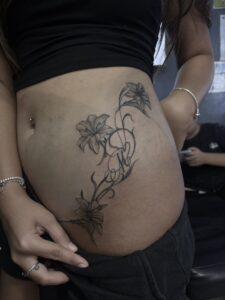 A delicate fine-line stomach tattoo for a woman, featuring a vertical flow of lilies and ornamental vines intertwined with a custom initial.