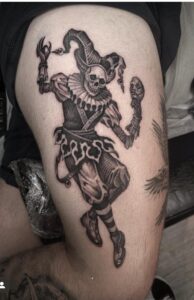 An illustrative black and grey thigh tattoo of a skeletal jester in full costume, wearing a ruffled collar and jester's hat while holding a theatrical mask.