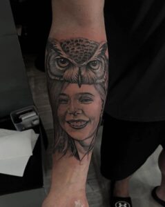 A realistic black and grey forearm tattoo featuring a portrait of a smiling young girl wearing an owl as a headdress, showcasing detailed feather textures and fine-line facial features.