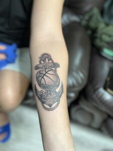 A custom black and grey forearm tattoo by Kurt Bayeta, featuring a detailed basketball integrated with an ornate anchor and nautical rope, showcasing crisp linework and smooth shading.