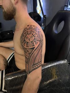 A man with a traditional tribal tattoo covering his right shoulder and arm, seated in a tattoo chair.