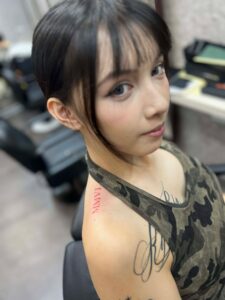 A woman with short, dark hair and bangs looks over her shoulder, showing a red Roman numeral tattoo "MMVI" on her upper shoulder, along with other script tattoos on her arm.