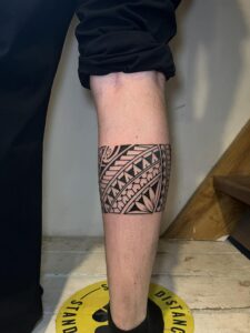 A bold Polynesian tribal leg band tattoo on the calf, featuring traditional geometric patterns, triangular motifs, and floral-inspired elements in solid black ink.