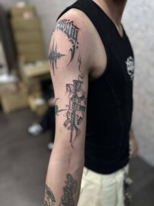 A black and grey illustrative tattoo of a rifle on a person's upper arm, featuring smoking barrel details and falling bullet casings.