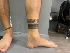 Close-up of a person's lower legs. The right leg features a detailed black tribal armband tattoo with geometric patterns and lines. The left ankle has a brown and gold woven bracelet.