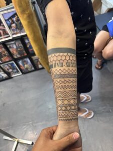 A forearm tattoo sleeve featuring a solid black band at the top, a honeycomb pattern with Roman numerals, and multiple rows of geometric tribal-style linework extending to the wrist.