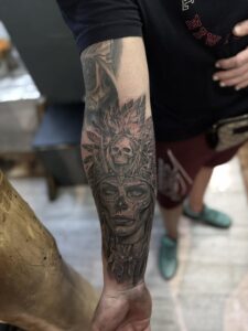 A detailed black and grey forearm tattoo of a woman’s face wearing an elaborate feather headdress with a central skull, featuring realistic shading and ornate jewelry details.