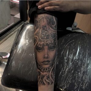 A realistic black and grey forearm tattoo of a woman's face wearing a decorative skull headdress, featuring soft shading and intricate ornamental details.