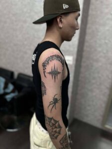 Side view of a blackwork patchwork arm sleeve featuring arched gothic lettering on the shoulder, a geometric star symbol, a spider, and an elbow spiderweb.