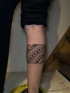 A bold Polynesian tribal leg band tattoo on the calf, featuring traditional geometric patterns, triangular motifs, and floral-inspired elements in solid black ink.