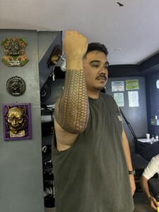A man with a mustache and a grey tank top raises his arm, proudly displaying a traditional tribal tattoo that covers his forearm and extends towards his elbow.