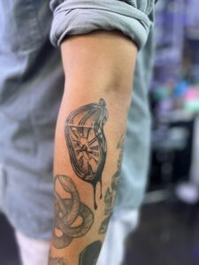 person's arm is visible, adorned with a black ink tattoo of a melting clock with Roman numerals.