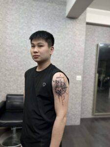 A man's upper arm tattoo featuring a realistic wolf head merged with a geometric compass, topped with Roman numerals in a fine-line black and grey style.