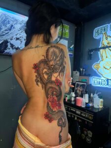 A large black and grey Japanese dragon back tattoo by Kurt Bayeta, featuring intricate scales, a dynamic serpentine flow, and vibrant pink cherry blossoms for a striking color contrast.