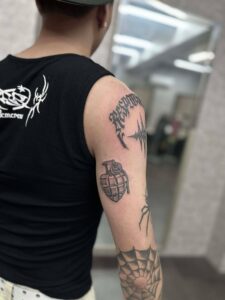 A patchwork tattoo collection on a man's arm, featuring a detailed grenade on the tricep, gothic script on the shoulder, and a spider design on the elbow.