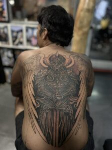 A large full-back tattoo of a winged warrior in ornate plate armor, holding a broadsword downward, featuring detailed shading and large feathered wings.